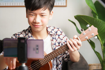 Online music lessons, live streaming concept. Handsome Asian teenager boy using his smartphone to broadcast or live his ukulele song on social media. Internet 5G, Lockdown, New normal, vlogger.