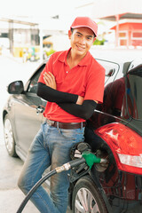 Asian gas station worker leaning against black car with happiness while green fuel nozzle filling high energy power fuel into black car tank, commercial service for benzine, diesel, gasohol, gasoline