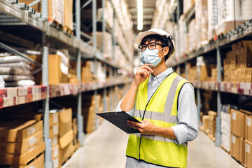 Portrait of asian engineer man in helmet in quarantine for coronavirus wearing protective mask order details checking goods and supplies on shelves with goods background in warehouse.logistic