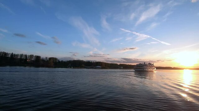 The passage of a large passenger ship along the river at sunset. Beautiful colorful landscape of the river against the background of the blue sky at sunset. Traveling by ship on the river