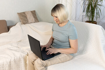 Smiling attractive adult woman with blonde hair sitting on comfy sofa at home working on laptop computer. FLowers on background. Serious lady.