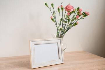 Portrait white picture frame mockup on wooden table. Modern ceramic vase with carnation. White wall background. Scandinavian interior. 