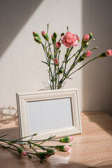 Portrait white picture frame mockup on wooden table. Modern ceramic vase with carnation. White wall...