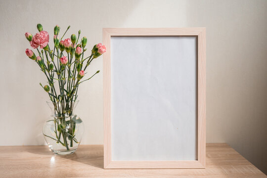 Portrait white picture frame mockup on wooden table. Modern ceramic vase with carnation. White wall background. Scandinavian interior. 