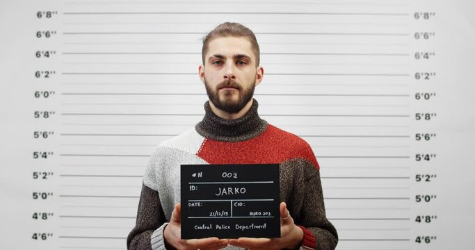 Portrait of handsome guy with beard holding sign for photo in police department. Crop view of young man with mustaches posing, raising head and looking to camera.Concept of criminal.