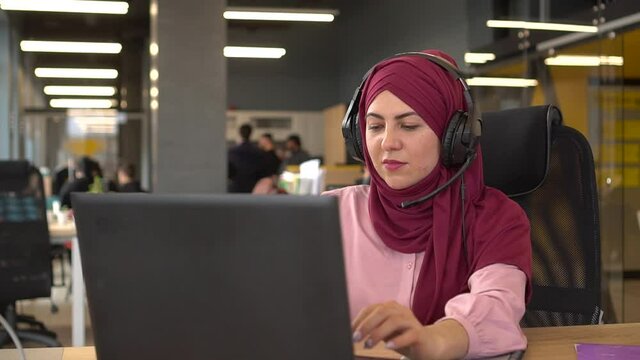 Friendly and smiling Muslim businesswoman with headphones and a microphone works in the office with a multi-racial team. Online video communication, customer support, remote working