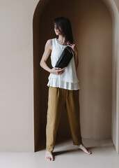 Black Leather Waist Bag For Women Small Leather Bum Bag. Young caucasian woman stands barefoot indoors. The woman is wearing brown pants, a white blouse and a black banana bag.