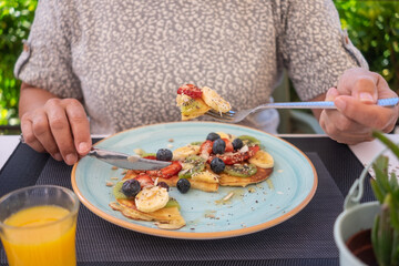 Mature woman eating healthy summer breakfast, classic american pancakes with banana, kiwi, fresh berry and honey