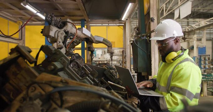 Professional industrial engineer African american man using computer laptop to control and programing automation robot arms machine in manufacturing industry. People and Innovation Technology concept.