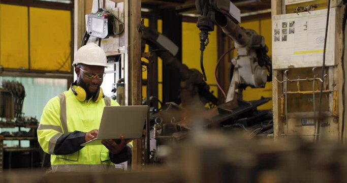 Professional industrial engineer African american man using computer laptop and to control and programing automation robot arms machine in manufacturing industry. Technology and innovation concept.