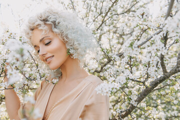 Portrait of a young blonde woman with curly hair at a  cherry tree with white flowers, smiling.