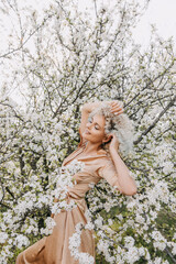 Young blonde woman with curly hair, smiling, at a cherry tree with white flowers on a spring day.