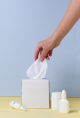 Vertical image of woman hand who taking paper napking for her stuffy nose, white bottle of nasal drops on the yellow desk against blue background