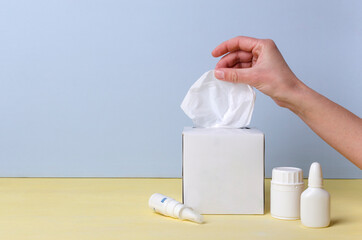 Closeup of woman hand, white container of paper napkins, bottle of vitamins and nasal drops on the desk against blue wall.Empty space