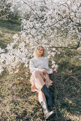 Young blonde woman with curly hair, wearing a sweater and rubber boots, lying under a cherry tree...