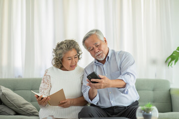 Happy Wellness Asian Elderly or Seniors couple holding mobile phone and book smile and laugh together on sofa in living room at home,Retirement lifestyle at home concept