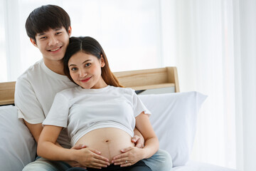 portrait of a lovely young couple dressed casually sitting on a bed together. A beautiful pregnant woman sitting and leaning on a handsome husband's chest happily. Family concept