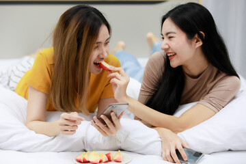 Obraz na płótnie Canvas Smiling Two Asian young women lovely couple repose on white bed and happy Feed apples. Funny women together on cozy. Concept In love homosexual lesbian lgbt, lgbtq, lgbtq+.
