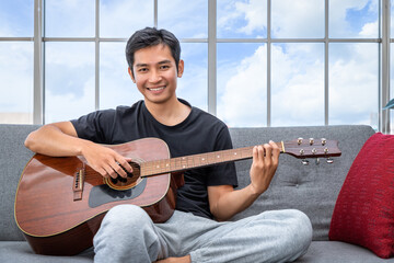 Handsome young Asian man playing acoustic guitar sitting on sofa in living room, looking at camera - Powered by Adobe