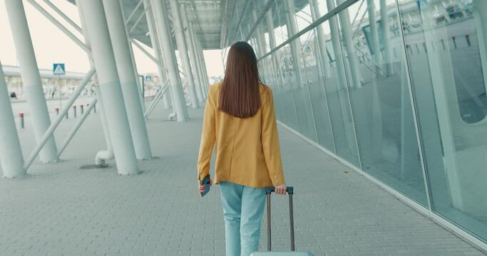 Back view of young woman in trendy yellow jacket and blue jeans going to airport with suitcase and smartphone in hands. Concept of people, technology and business trip. 