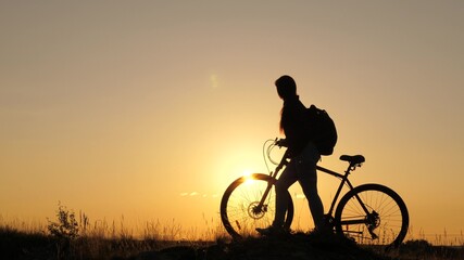 Fototapeta na wymiar A free girl travels on a bike, rests, looks at the sunset and enjoys the sun. Adventure and travel concept. A healthy young tourist walks with bicycles across the field, enjoying nature, fresh air.