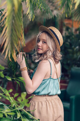 Positive blonde teenage girl in a straw hat walks among the palm trees, touches the leaves, rear view.Concept of closeness to nature.Eco,urban jungle,biophilia concept.Selective focus.