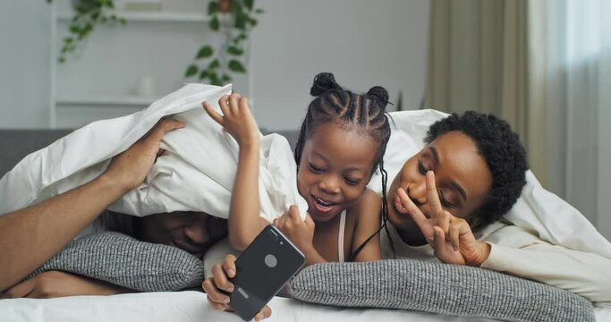 Funny Afro american happy ethnic black family lying on sofa covered with blanket, little african daughter child girl taking photo shoot video on mobile phone camera, mom posing, dad hiding laughing