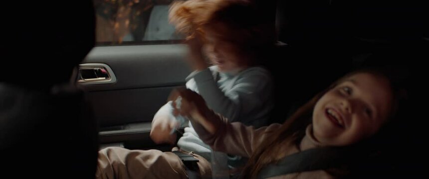 Cute little siblings brother and sister singing and going crazy on a back seat of a modern SUV while riding through neighborhood. Shot with 2x anamorphic lens