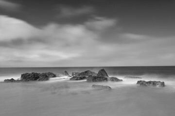 Naklejka na ściany i meble Long exposure photo black and white of waves breaking on rocks on the shoreline with a sky with clouds. Conil de la Frontera, Spain