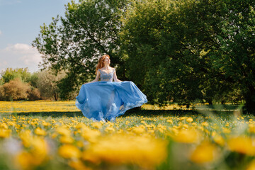 A young cheerful girl runs in a blue long dress, across a field with yellow meadow flowers. A summer lawn with dandelions.