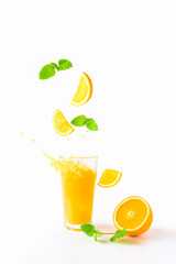 Pieces of yellow orange together with fresh lemon balm leaves fall into a glass of freshly squeezed orange juice. Juice splashes. Flying fruits. Half an orange next to the glass. White background.