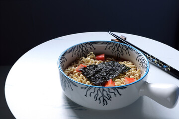 instant noodle with crab sticks and natural seawood cooked by olive oil in the nice white bowl is on white table with black chopsticks in the dark room during breakfast