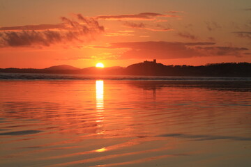 Obraz premium Setting sun drops to the horizon, silouetting Criccieth Castle, and casts reflections across the rippled sand at Black Rock Beach (landscape).