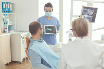Obraz premium Stomatologist nurse showing tooth x-ray to sick patient explaining treatment using tablet working in stomatology hospital office. Senior doctor examining toothache while man sitting on dental chair