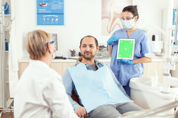 Obraz premium Stomatology nurse pointing her hand on mock up green screen chroma key tablet with isolated display. Sick man patient sitting on dental chair in stomatology office waiting for toothache treatment
