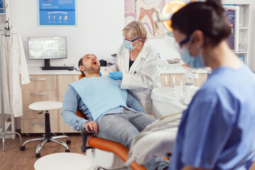 Obraz premium Sick patient sitting on dental chair with open mouth while senior dentist woman analyzing health teeth before medical surgery. Stomatological nurse preparing stomatology tools