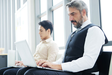 Fototapeta premium Serious Senior Caucasian businessman is working with laptop in workspace at office. He sitting distance from asian staff. Technology for business and New normal lifestyle concept.