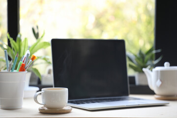 White coffee cup and laptop on table and plant pot in front of window
