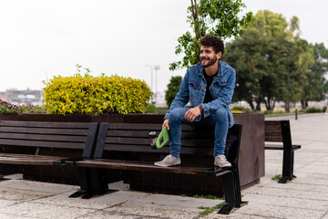 Fototapeta premium Young handsome man with curly hair and a beard wearing denim sitting on a bench with a skateboard in an empty public park
