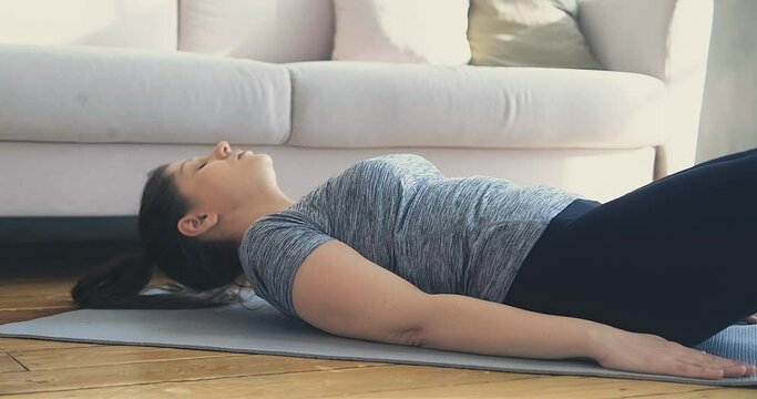 Athletic young woman in tracksuit does crunches and leg lifts lying on grey mat in light living room close view slow motion