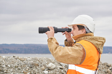 mining or road engineer using a spotting scope against the background of a riverbank quarry