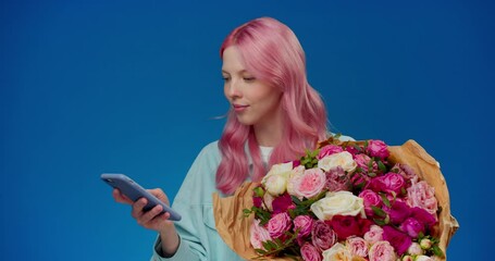 Happy young woman with pink hair holding bouquet of flowers types the message phone thanks for gift and smelling roses on red background. Cute girl happy with flower delivery