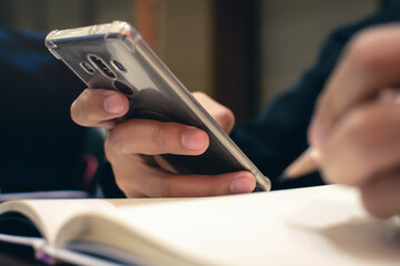 Close up man holding and using mobile phone with another hand holding the pen on notebook.