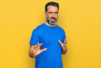 Middle aged man with beard wearing casual blue t shirt disgusted expression, displeased and fearful doing disgust face because aversion reaction. with hands raised