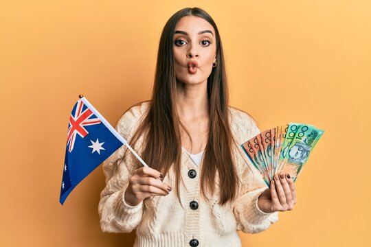 Beautiful brunette young woman holding australian flag and dollars making fish face with mouth and squinting eyes, crazy and comical.