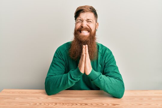 Young irish redhead man wearing casual clothes sitting on the table praying with hands together asking for forgiveness smiling confident.