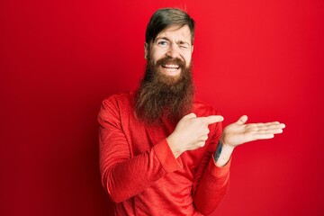 Redhead man with long beard pointing open palm with finger winking looking at the camera with sexy expression, cheerful and happy face.