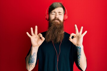 Redhead man with long beard listening to music using headphones relax and smiling with eyes closed doing meditation gesture with fingers. yoga concept.
