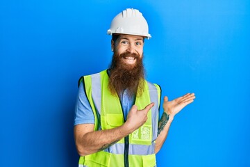 Redhead man with long beard wearing safety helmet and reflective jacket amazed and smiling to the camera while presenting with hand and pointing with finger.