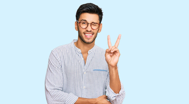 Young handsome man wearing casual clothes and glasses smiling with happy face winking at the camera doing victory sign. number two.